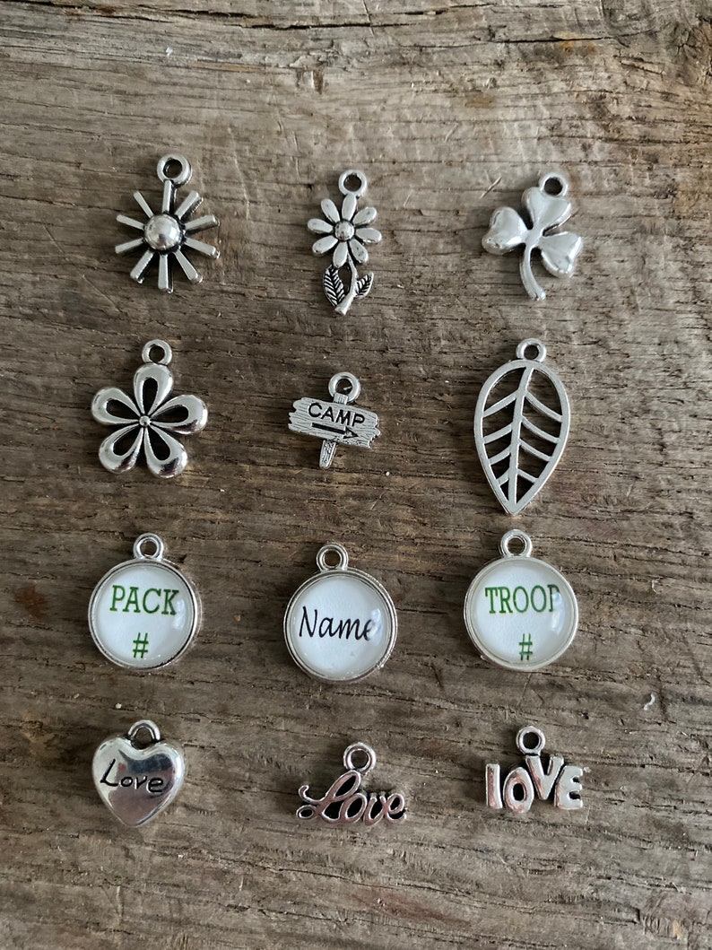 Peut inclure: Une collection de breloques argent&eacute;es de diff&eacute;rentes formes, dont des fleurs, un tr&egrave;fle, une feuille et des c&oelig;urs. Certaines breloques portent les mots "Pack", "Name", "Troop" et "Love". Les breloques sont pr&eacute;sent&eacute;es sur une surface en bois.