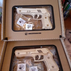 May include: Two cardboard boxes with clear plastic windows showing wooden toy guns. The top box has a gun labeled "J.D." and the bottom box has a gun labeled "H.D.". Both boxes have a small logo with a cartoon animal on them.