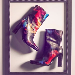 May include: A pair of black leather boots with colorful abstract paint splatter designs. The boots have a high heel and a pointed toe.
