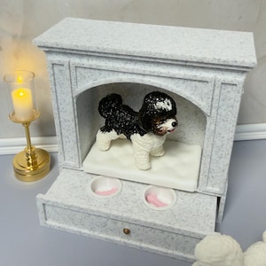 May include: A miniature dog figurine with black and white fur stands inside a decorative, speckled gray dog house. The dog house has a drawer with two pink bowls. A gold candle holder with a lit candle is to the left.