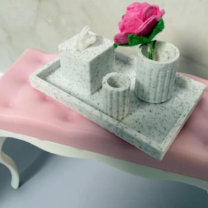 May include: A decorative vanity set with a pink surface and a white table. The set includes a tray, a tissue box, a small cup, and a larger cup with a pink rose. The items have a speckled pattern.