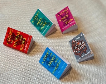 May include: Five paperback books with colorful covers and the title "A Court of" on each. The books are stacked in a pyramid shape on a beige fabric surface. The titles are "A Court of Thorns and Roses", "A Court of Mist and Fury", "A Court of Wings and Ruin", "A Court of Frost and Starlight", and "A Court of Silver Flames".