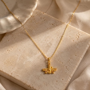 May include: A gold-colored necklace with a pendant featuring a detailed, stylized design. The pendant is suspended from a delicate chain. The necklace is displayed on a textured, light-colored surface, with a soft, white fabric in the background.