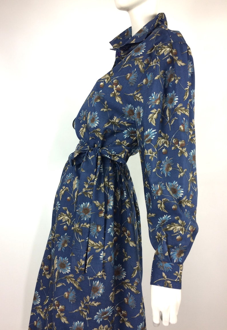 Puede incluir: Un vestido azul de manga larga con estampado floral. El vestido tiene cuello, cierre de botones en la parte delantera y un cintur&oacute;n de lazo en la cintura.