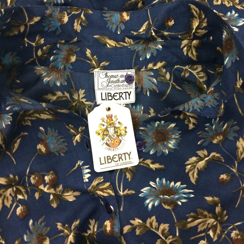 Puede incluir: Una camisa de algod&oacute;n azul con estampado floral de flores marrones y azules. La camisa tiene cuello abotonado y una etiqueta que dice "Thomas and Jonathan of Canterbury" y "Liberty, Made in England".