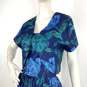 K&ouml;nnte beinhalten: Ein blaues und gr&uuml;nes Blumenkleid mit einer passenden kurzen Jacke. Das Kleid hat ein tailliertes Oberteil und einen vollen Rock.