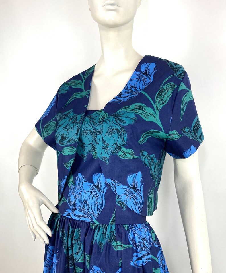 K&ouml;nnte beinhalten: Ein blaues und gr&uuml;nes Blumenkleid mit einer passenden kurzen Jacke. Das Kleid hat ein tailliertes Oberteil und einen vollen Rock.