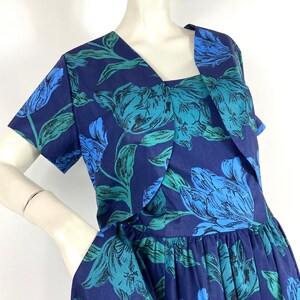 K&ouml;nnte beinhalten: Ein marineblaues Kleid mit einem Blumendruck aus blauen und gr&uuml;nen Tulpen. Das Kleid hat einen V-Ausschnitt und kurze &Auml;rmel. Der Rock ist in der Taille gerafft.