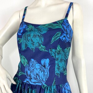 K&ouml;nnte beinhalten: Ein &auml;rmelloses, marineblaues Kleid mit einem Blumendruck mit gro&szlig;en blauen und gr&uuml;nen Blumen. Das Kleid hat einen figurbetonten Oberteil und einen flie&szlig;enden Rock.