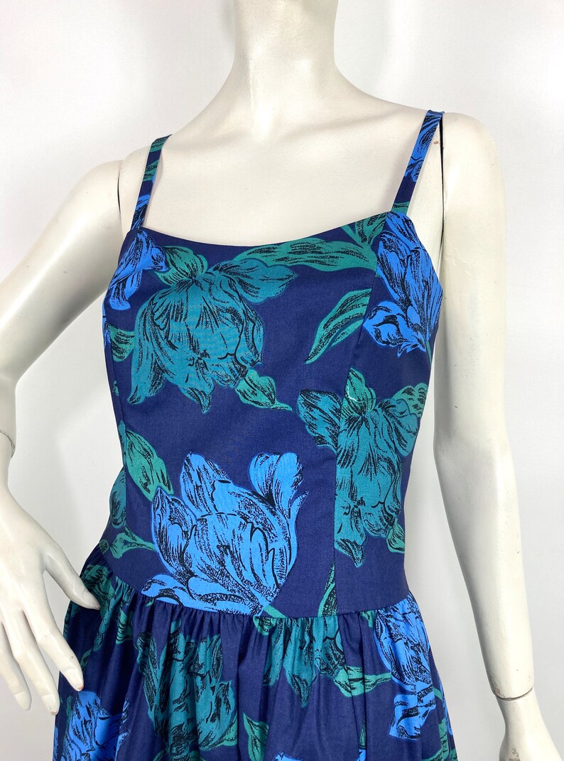 K&ouml;nnte beinhalten: Ein &auml;rmelloses, marineblaues Kleid mit einem Blumendruck mit gro&szlig;en blauen und gr&uuml;nen Blumen. Das Kleid hat einen figurbetonten Oberteil und einen flie&szlig;enden Rock.