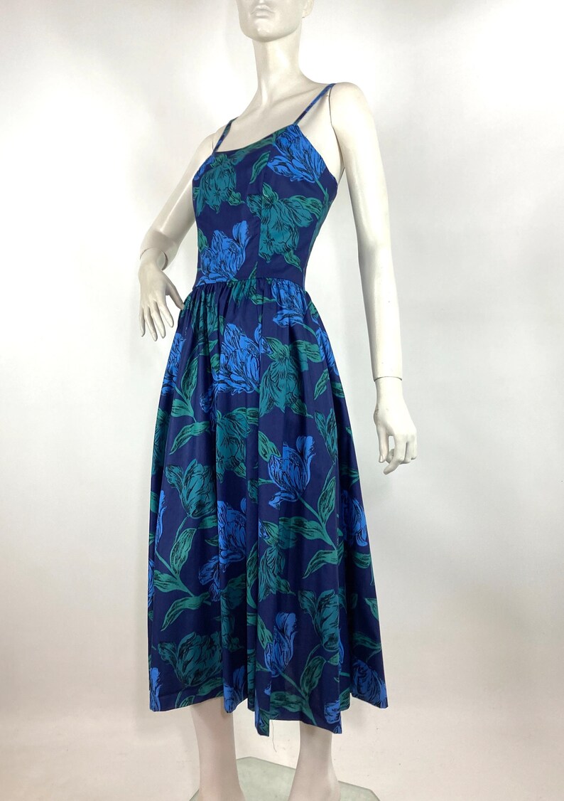 K&ouml;nnte beinhalten: Ein blaues Midikleid mit einem Blumendruck mit gr&uuml;nen Bl&auml;ttern und blauen Bl&uuml;ten. Das Kleid hat einen taillierten Oberteil und einen weiten Rock.