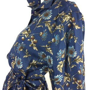 Puede incluir: Un vestido azul de manga larga con estampado floral de tallos marrones y flores azules. El vestido tiene un cierre de botones en la parte delantera y un lazo en la cintura.