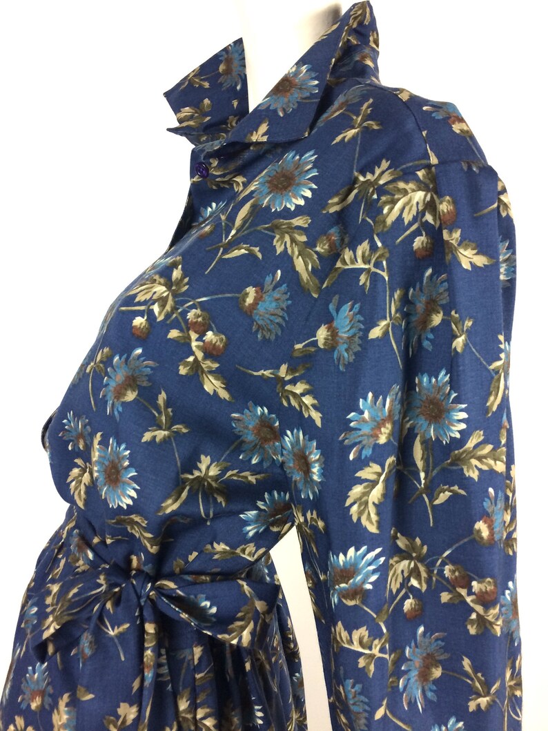 Puede incluir: Un vestido azul de manga larga con estampado floral de tallos marrones y flores azules. El vestido tiene un cierre de botones en la parte delantera y un lazo en la cintura.