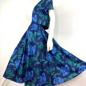 K&ouml;nnte beinhalten: Ein blaues und gr&uuml;nes Blumenkleid mit einem taillierten Oberteil und einem weiten Rock. Das Kleid hat kurze &Auml;rmel und einen V-Ausschnitt.