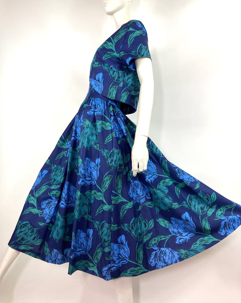 K&ouml;nnte beinhalten: Ein blaues und gr&uuml;nes Blumenkleid mit einem taillierten Oberteil und einem weiten Rock. Das Kleid hat kurze &Auml;rmel und einen V-Ausschnitt.