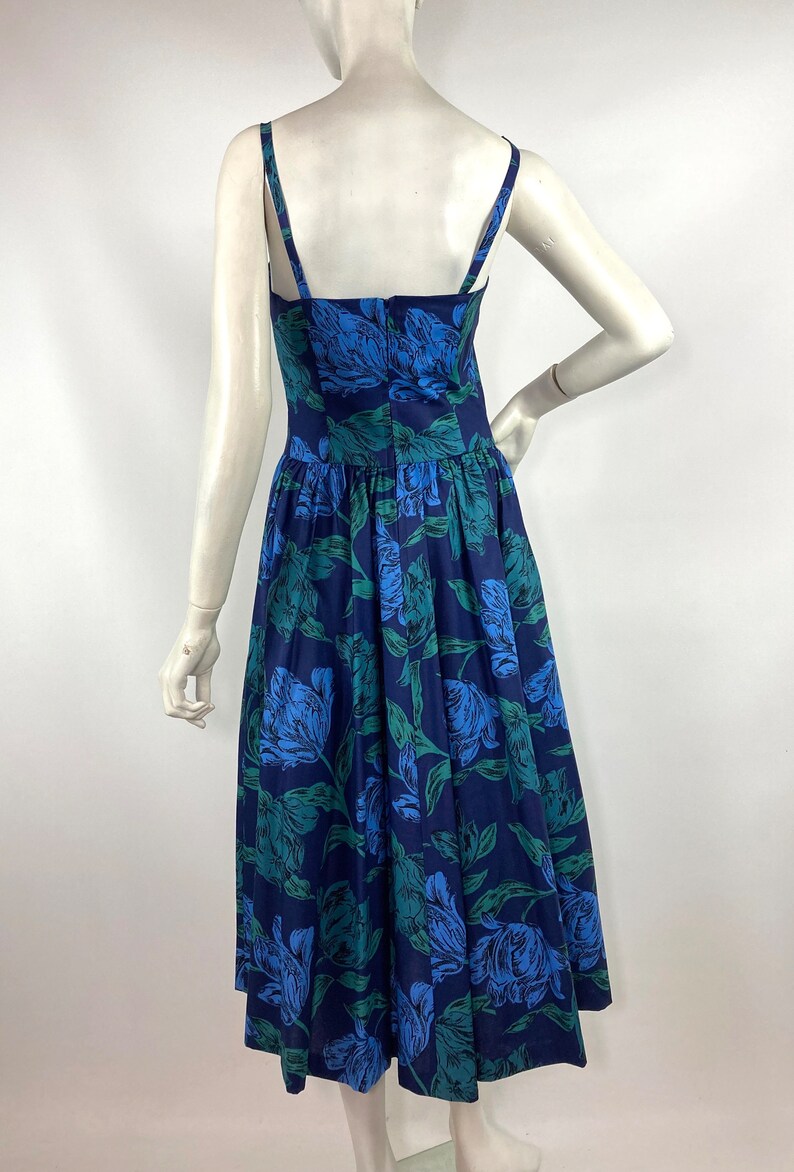 K&ouml;nnte beinhalten: Ein blaues und gr&uuml;nes Blumenmuster-Midi-Kleid mit d&uuml;nnen Tr&auml;gern. Das Kleid hat ein tailliertes Oberteil und einen vollen Rock.