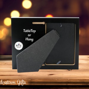 May include: Black picture frame with a tabletop or hang design. The back of the frame is visible, showing a stand and hanging hardware. The frame has a logo that says "BTG Budderstown Gifts".
