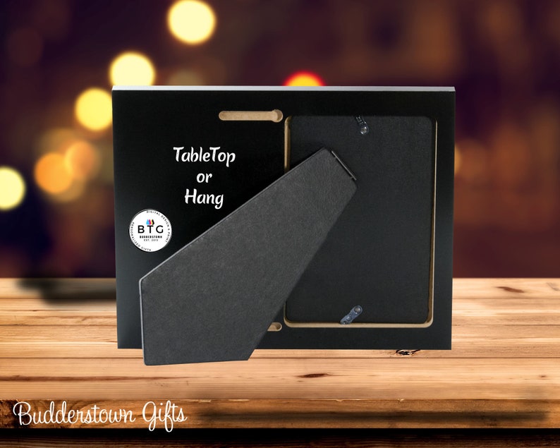 May include: Black picture frame with a tabletop or hang design. The back of the frame is visible, showing a stand and hanging hardware. The frame has a logo that says "BTG Budderstown Gifts".