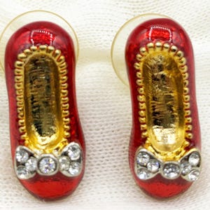 May include: A pair of red ballet slipper earrings with gold-colored interiors and trim. Each slipper has a bow adorned with clear rhinestones. The earrings have clear, round posts.