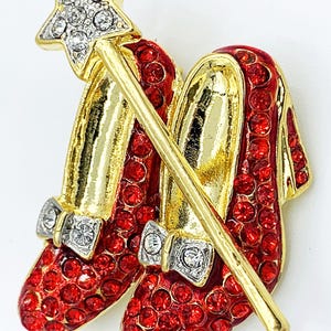 May include: A gold-tone brooch featuring a pair of red ruby-encrusted shoes with a gold wand and a star-shaped top. The wand is adorned with clear rhinestones.