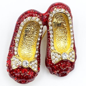 May include: A red and gold brooch in the shape of ballet slippers. The slippers are adorned with clear rhinestones and gold bows. The interior of the slippers is gold. The brooch has a pin on the back.