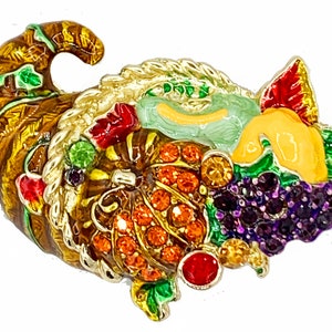 May include: A cornucopia filled with colorful fruits and vegetables, including grapes, pumpkins, and apples. The cornucopia is made of metal and is decorated with rhinestones.