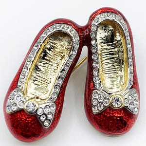 May include: A red enamel shoe brooch with a gold interior and silver trim. The brooch features two shoes connected at the heels, each adorned with a bow and sparkling rhinestones. A gold pin is attached to the back.