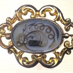 May include: A gold-toned brooch with an oval-shaped glass cameo featuring a design of hair woven into a floral pattern. The cameo is set in a decorative frame with scrollwork.