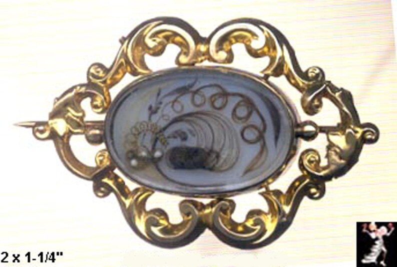 May include: A gold-toned brooch with an oval-shaped glass cameo featuring a design of hair woven into a floral pattern. The cameo is set in a decorative frame with scrollwork.