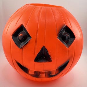 May include: A bright orange plastic pumpkin bucket featuring a carved jack-o'-lantern face with black painted accents around the diamond-shaped eyes, triangular nose, and wide, toothy grin. The surface has vertical ridges and a hollow top opening.