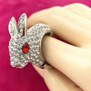 May include: A silver-toned ring featuring a rabbit design. The rabbit's head and body are encrusted with clear crystals, with a red crystal eye. The ring is a statement piece, suitable for adding a touch of whimsy to any outfit.