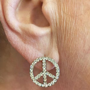 Peut inclure: Gros plan d'une boucle d'oreille à clou en forme de signe de paix argenté. Le symbole de la paix est souligné de petits cristaux transparents. La boucle d'oreille est placée sur le fond d'une oreille et de la peau.