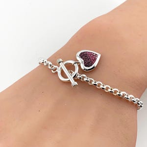 May include: A silver-toned chain bracelet featuring a toggle clasp and a heart-shaped charm. The charm is encrusted with small, dark pink or magenta crystals that create a textured, sparkling surface against the polished silver metal finish.