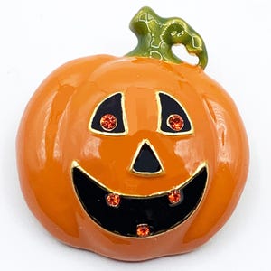 May include: Orange pumpkin brooch with a smiling face, black eyes, and a black mouth. The eyes and mouth have orange crystal accents. The stem is green. A festive accessory for Halloween.