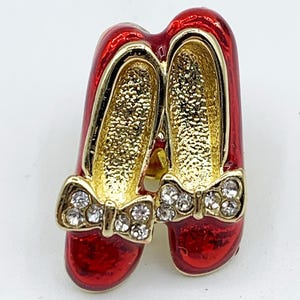 May include: A red and gold-toned brooch in the shape of a pair of ruby slippers. The slippers have gold interiors and are adorned with rhinestone-studded bows. The brooch has a glossy finish and a gold border.