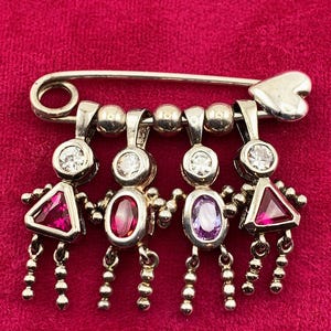 May include: Silver brooch with four figures holding hands, each with a gemstone head and body. The figures have dangling bead accents. The brooch has a safety pin closure and a heart-shaped detail. Set against a red background.