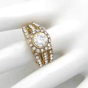 May include: A gold ring with a large clear stone surrounded by smaller clear stones. The ring has three bands of smaller stones.