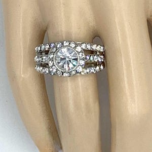May include: A silver ring with a large, clear, round center stone, surrounded by smaller, clear stones. The ring has three bands of stones, creating a layered effect. The ring is on a light-toned hand.