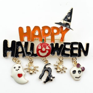 May include: A gold-tone metal brooch featuring the text HAPPY HALLOWEEN in orange and black enamel. A black witch hat sits atop the text. Five charms hang from the bottom: a white ghost with a red heart, two gold spiders, a black cat, and a skull.