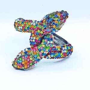 May include: A silver-toned hinged bracelet featuring a butterfly design. The butterfly wings and body are encrusted with a multitude of colorful, sparkling rhinestones in various shades of pink, blue, green, yellow, and orange. The bracelet is set against a white background.