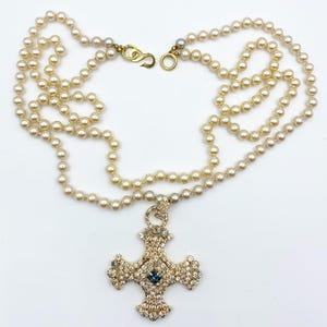 May include: A cream-colored pearl necklace with a gold clasp and a large, ornate cross pendant. The cross is encrusted with clear and blue crystals, creating a sparkling effect. The necklace is displayed on a white background.