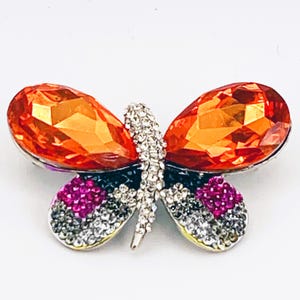 May include: A butterfly brooch featuring large orange gemstone wings. The body is silver with clear and pink crystals. The wings are a vibrant orange colour, set against a white background.