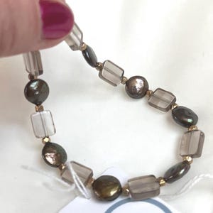 May include: A beaded bracelet featuring alternating smoky quartz rectangular beads and dark, iridescent, round and oval pearls. The bracelet is strung with gold-colored spacers and is held up by a hand with pink nail polish.