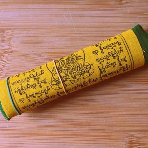 Puede incluir: Una bandera de oraci&oacute;n amarilla con texto negro y borde verde est&aacute; enrollada y asegurada con una banda el&aacute;stica. El texto en la bandera incluye las palabras "བཛགུརུཔདྨསིདྡྷིཧཱུྃ།ཨཾ" y "ཨཱཿ ཧཱུྃབཛྲགུརུཔདྨསི".