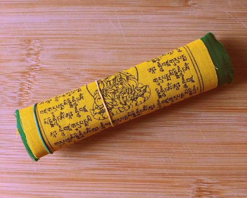 Puede incluir: Una bandera de oraci&oacute;n amarilla con texto negro y borde verde est&aacute; enrollada y asegurada con una banda el&aacute;stica. El texto en la bandera incluye las palabras "བཛགུརུཔདྨསིདྡྷིཧཱུྃ།ཨཾ" y "ཨཱཿ ཧཱུྃབཛྲགུརུཔདྨསི".