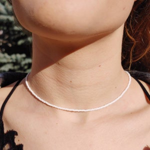May include: A simple white beaded choker necklace. The necklace is made of small, white beads that are strung together on a thin cord. The necklace is worn by a person with a light complexion.