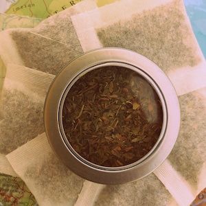 May include: A close-up of a round metal tin with a clear lid, filled with loose leaf green tea. The tin is surrounded by several white tea bags.
