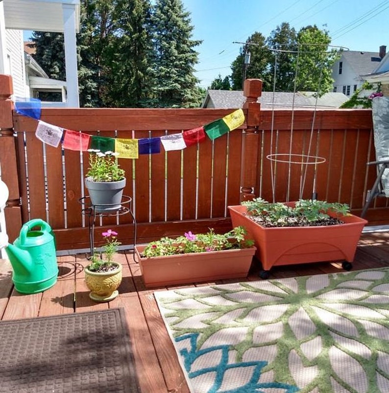 Puede incluir: Una terraza de madera con una valla de madera marr&oacute;n y una alfombra verde. Hay varias plantas en macetas, incluyendo una maceta grande de terracota con una planta creciendo dentro. Tambi&eacute;n hay banderas de oraci&oacute;n coloridas colgadas de la valla.