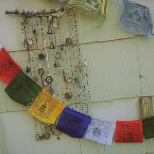 Puede incluir: Una hilera de banderas de oraci&oacute;n coloridas con mantras budistas tibetanos cuelga de una varilla de madera. Las banderas son rojas, verdes, amarillas, azules y blancas. Una cortina de cuentas con abalorios de metal cuelga en el fondo.