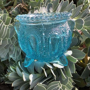 May include: A bright turquoise glass bowl featuring a textured, pebbled surface with a beaded rim and three short, sturdy feet. The bowl has a repeating arched pattern embossed around its exterior, creating a decorative, translucent appearance.